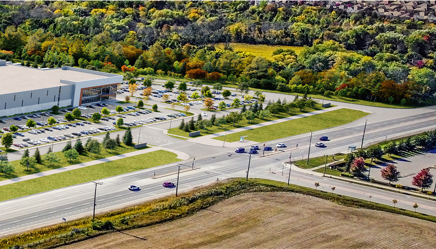 Aerial view of a commercial building with a landscaped parking lot, adjacent roadway, and surrounding fields and forested areas.
