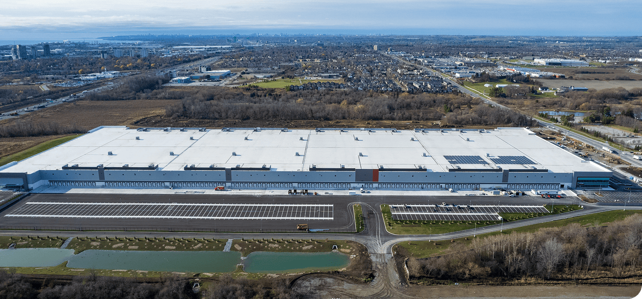 A wide aerial view of a large industrial distribution facility with a white roof, solar panels, stormwater ponds, and surrounding open land.