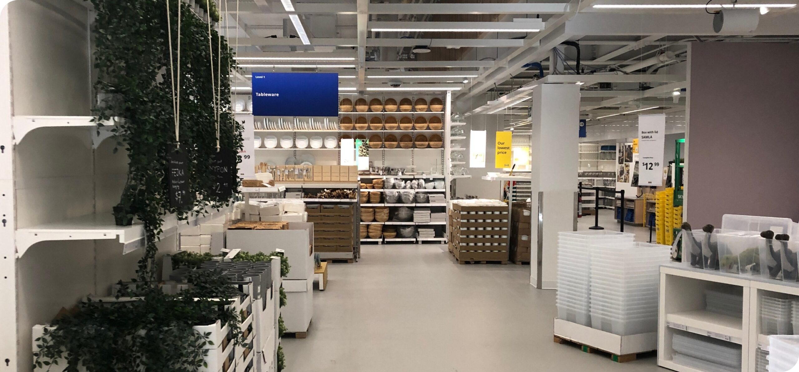 IKEA store interior with shelving units, storage products, and greenery displayed along wide aisles.