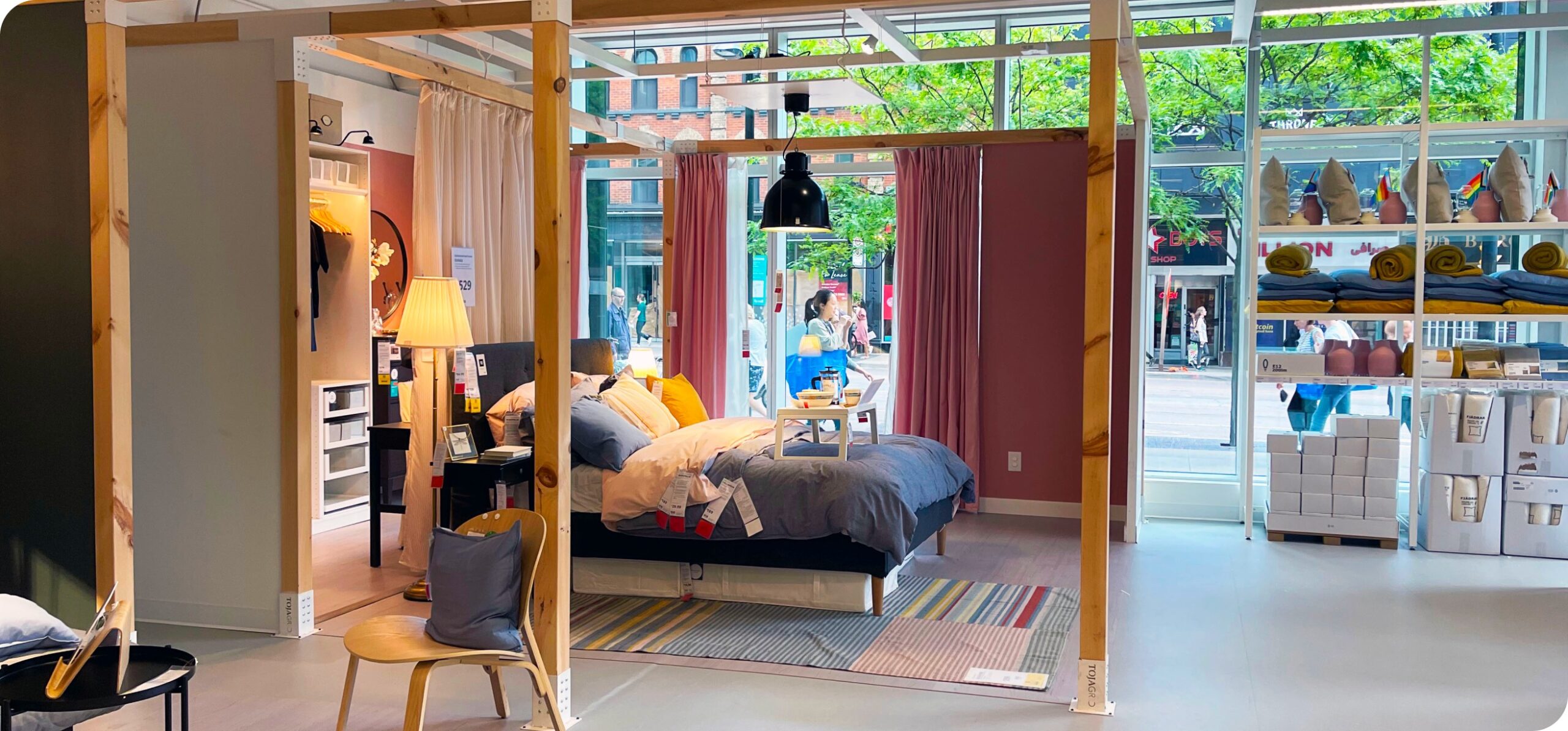 IKEA showroom bedroom display featuring a bed, lighting, soft furnishings, and windowed street-level backdrop.