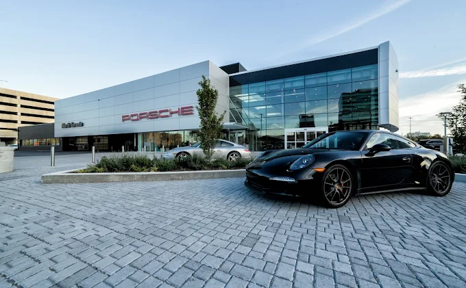 Modern Porsche dealership with a glass showroom and a black Porsche sports car parked in front on a stone-paved driveway.