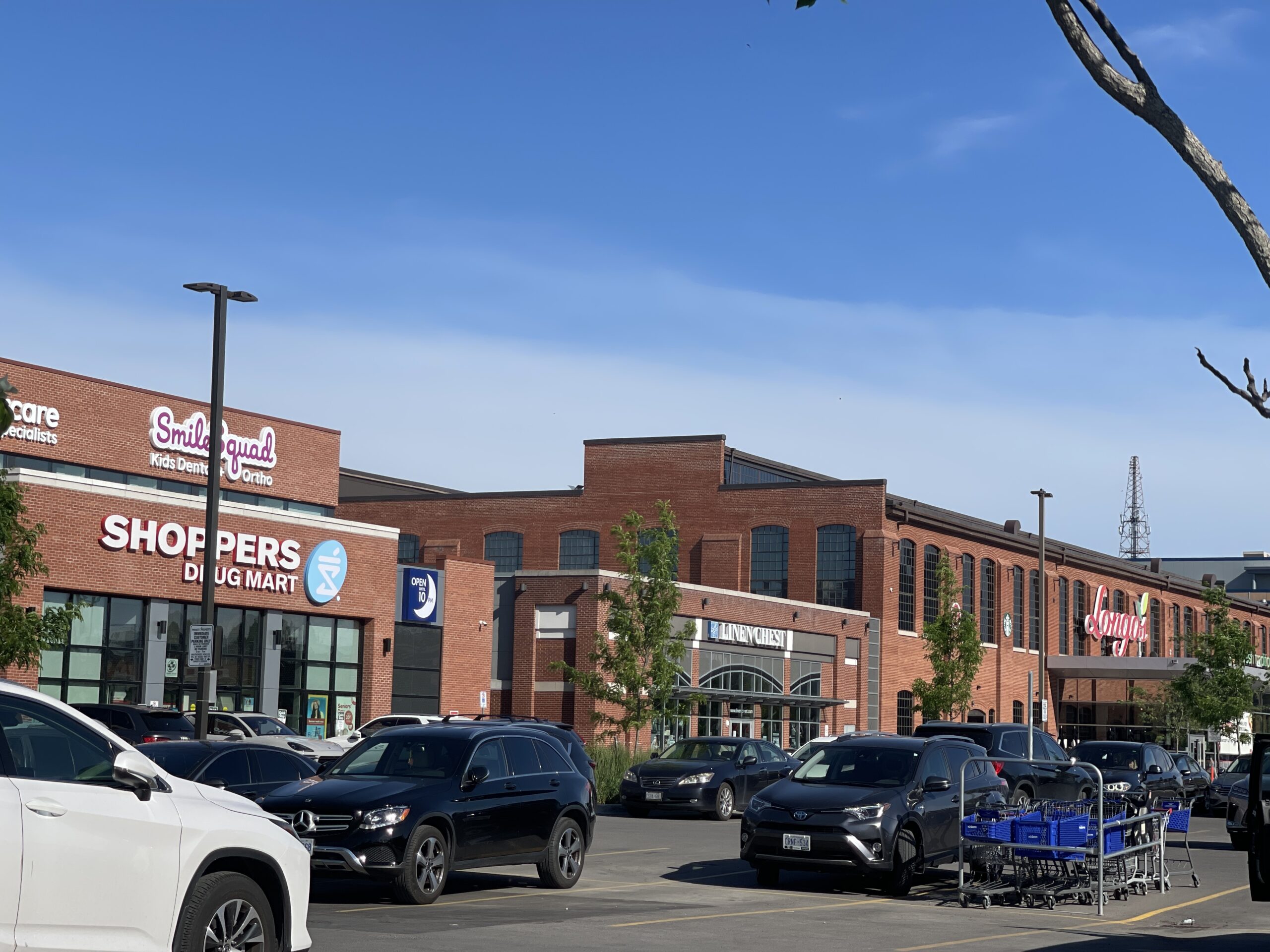 Street-level view of a retail plaza with brick façades, storefront signage, and a busy parking lot.