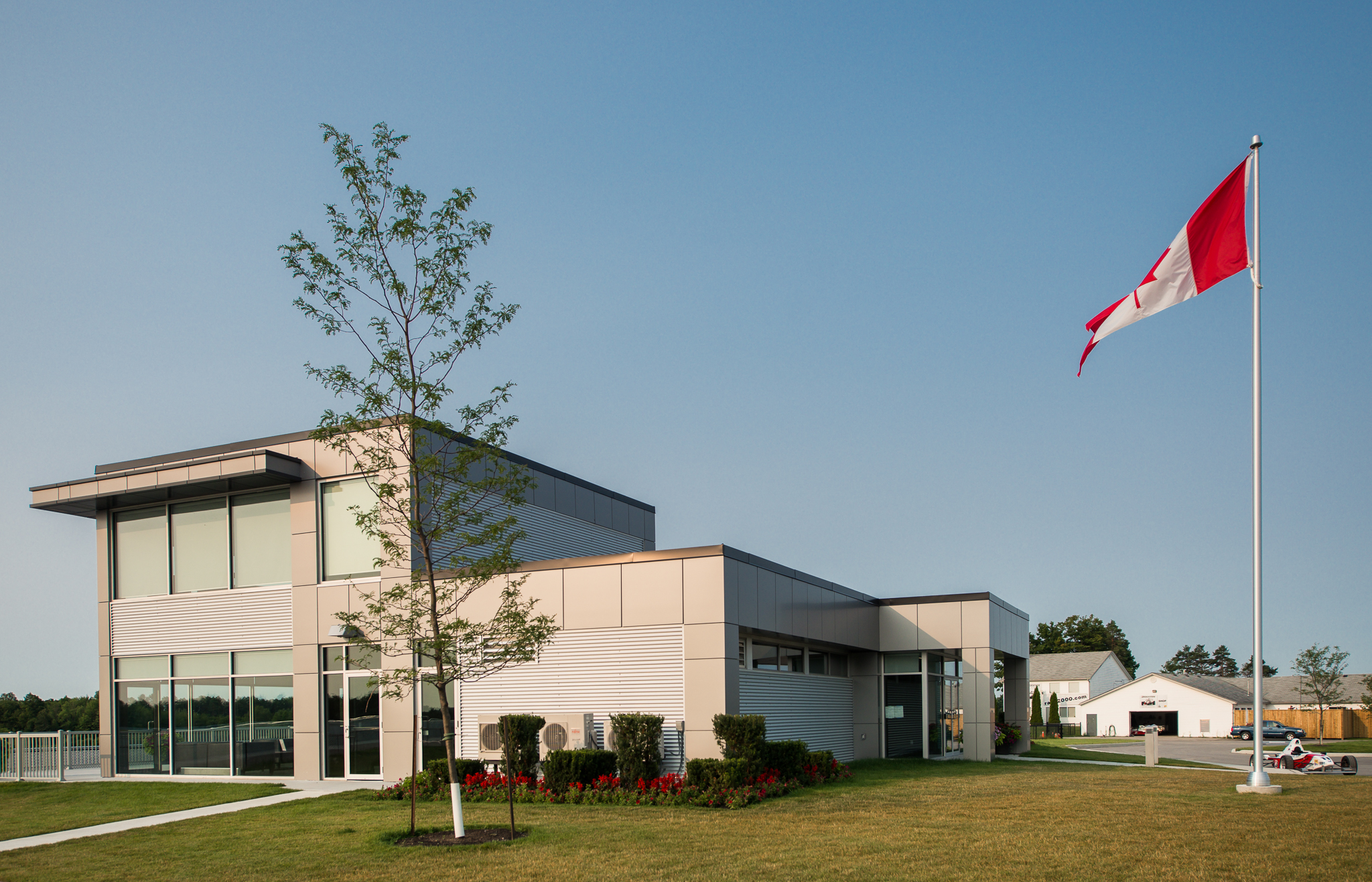 A modern building with large windows and a Canadian flag flying on a tall pole, surrounded by landscaped greenery.