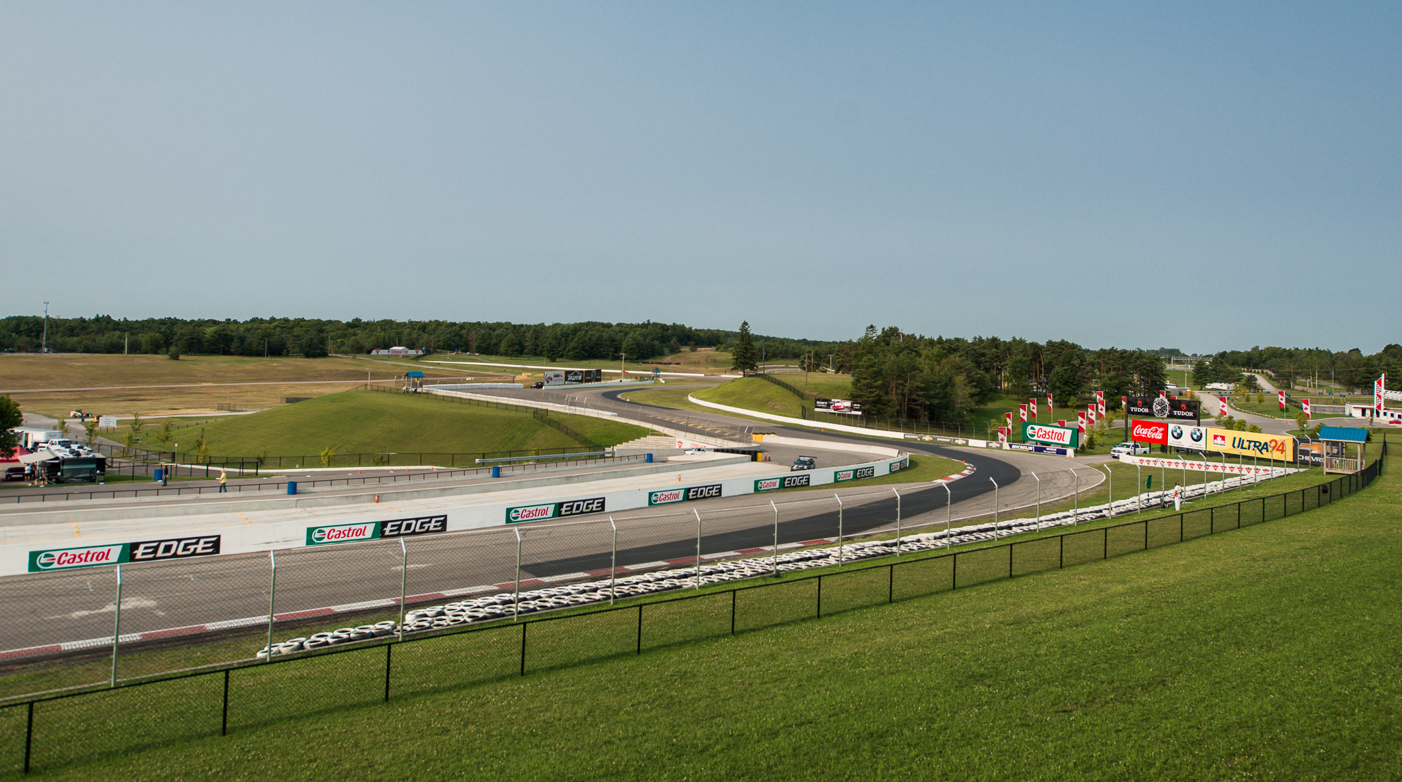 View of a motorsport racetrack with multiple turns, bordered by grassy hills and safety fencing.