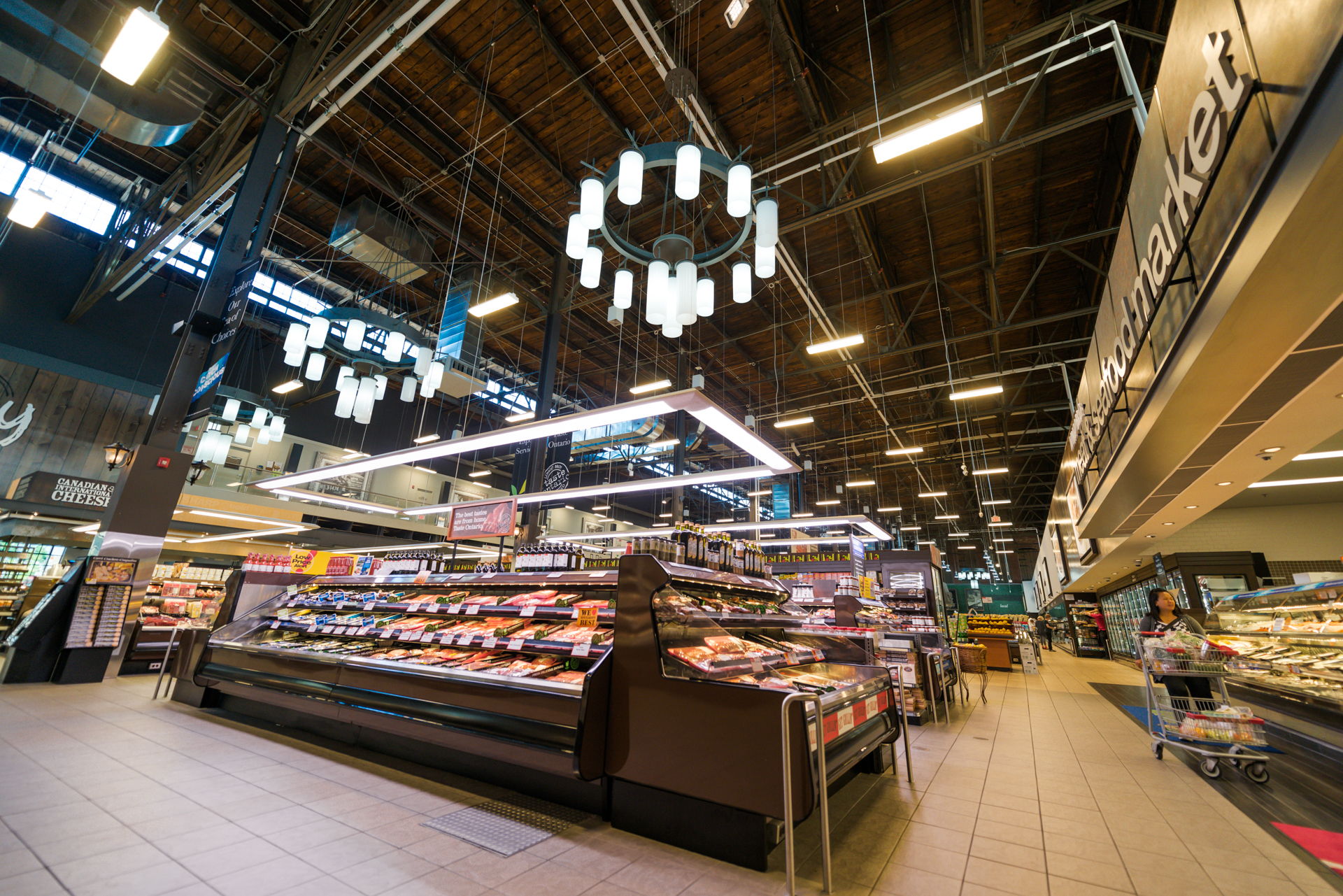 Grocery store interior with prepared-food displays, exposed wood ceiling, and modern lighting fixtures.
