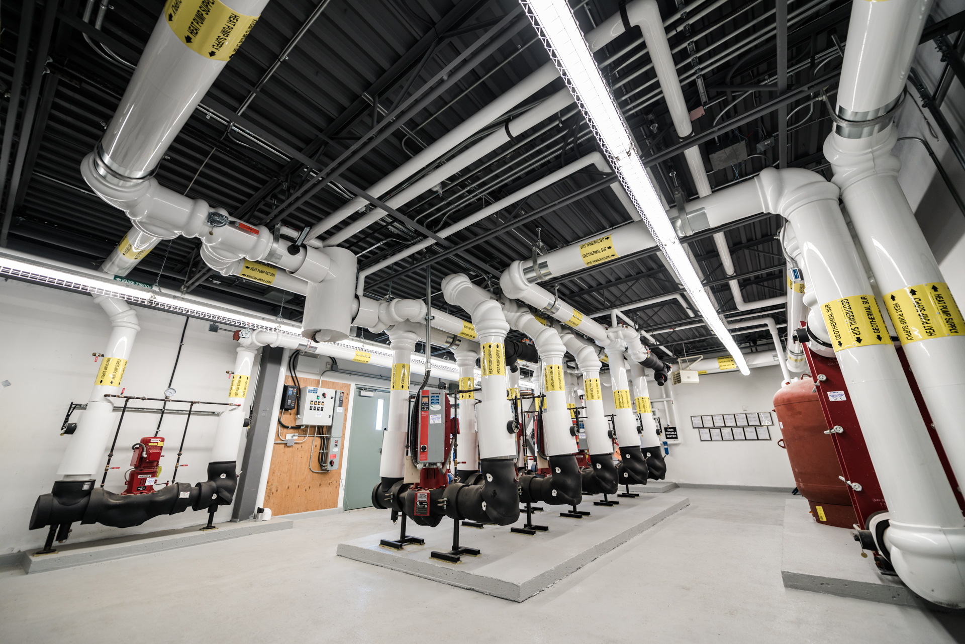 Mechanical room containing large white HVAC piping, pumps, and labelled utility lines.