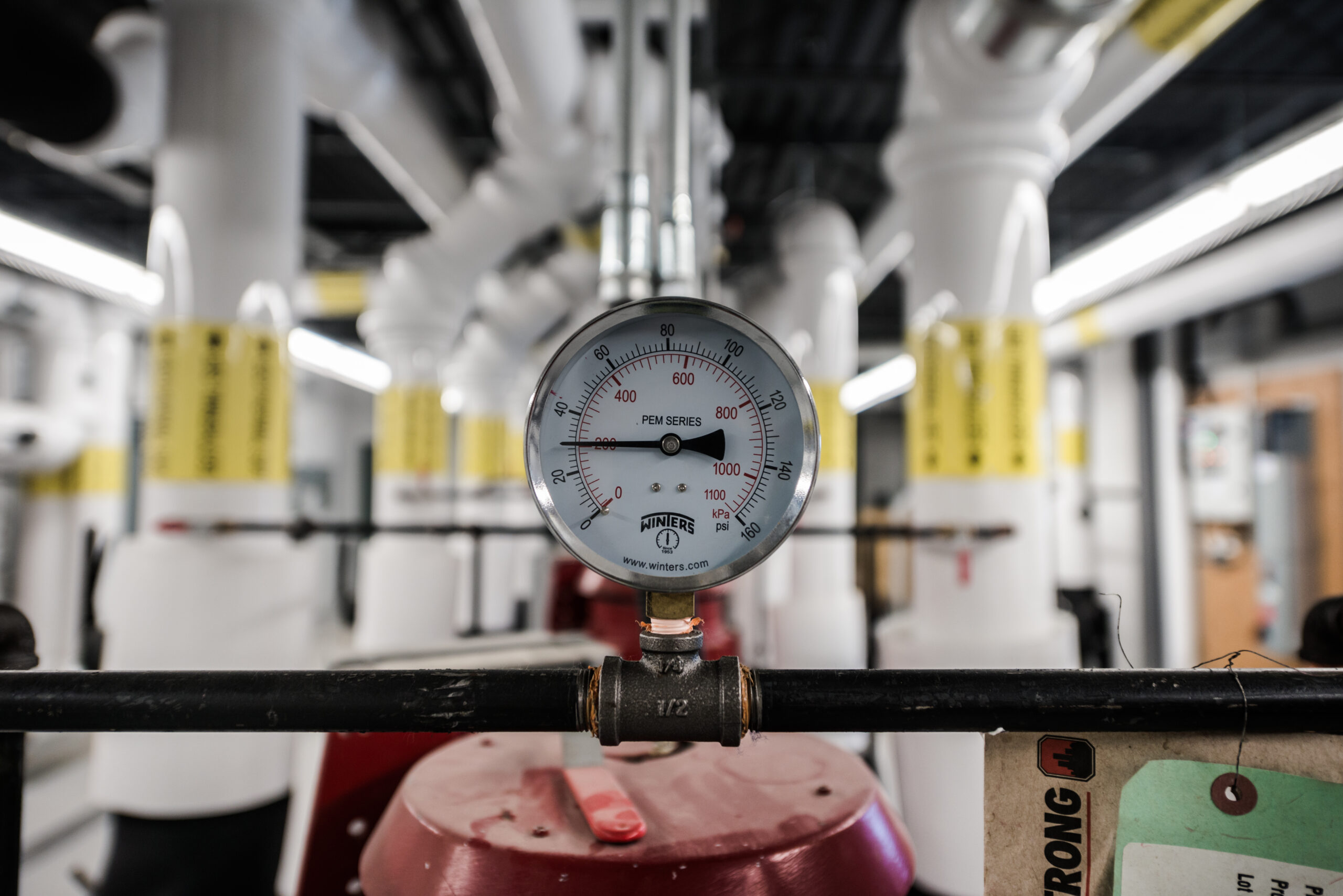 Close-up of a pressure gauge mounted on a pipe in a mechanical room, with insulated piping and equipment blurred in the background.