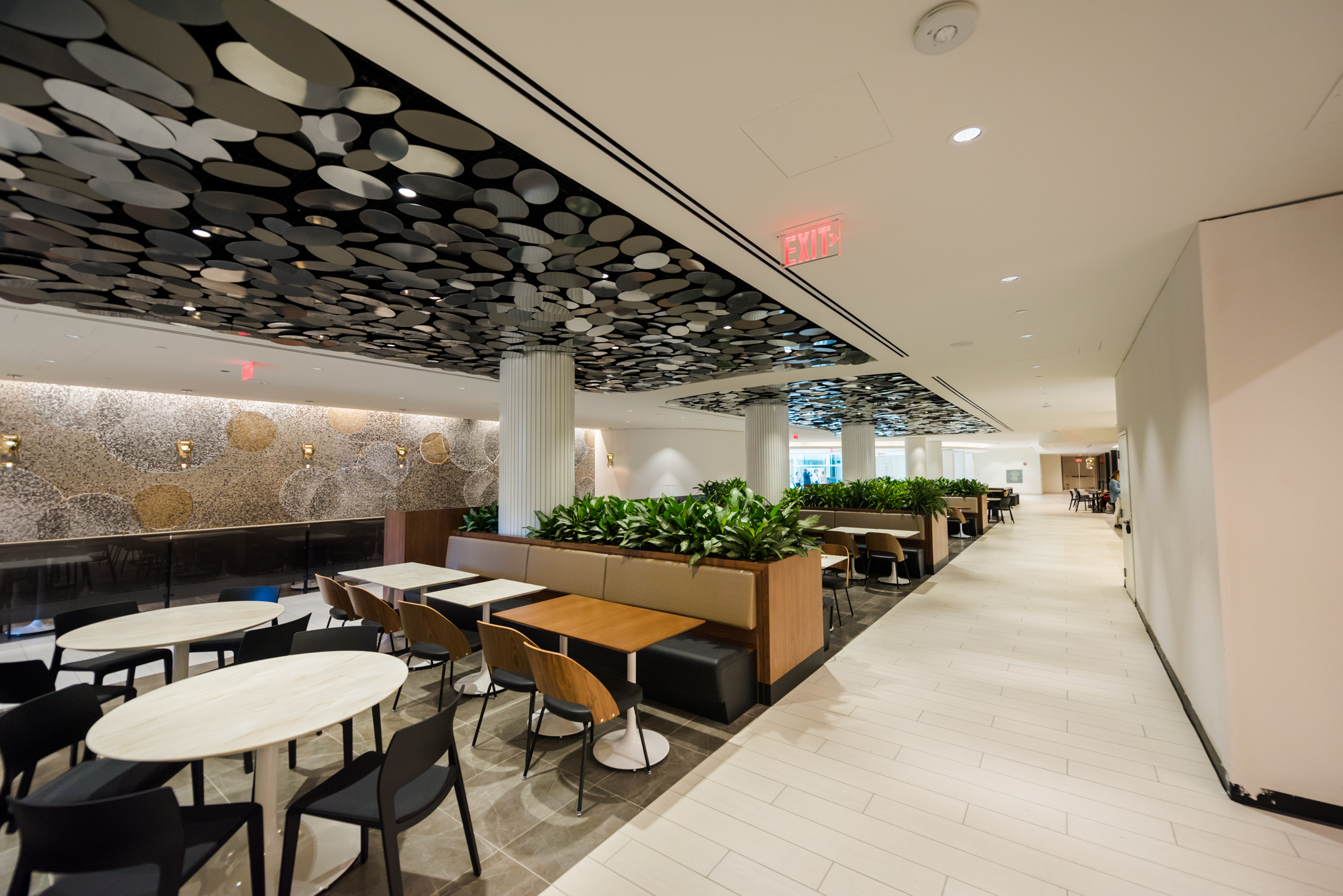 Modern cafeteria-style seating area with decorative ceiling panels, greenery dividers, and contemporary furniture.