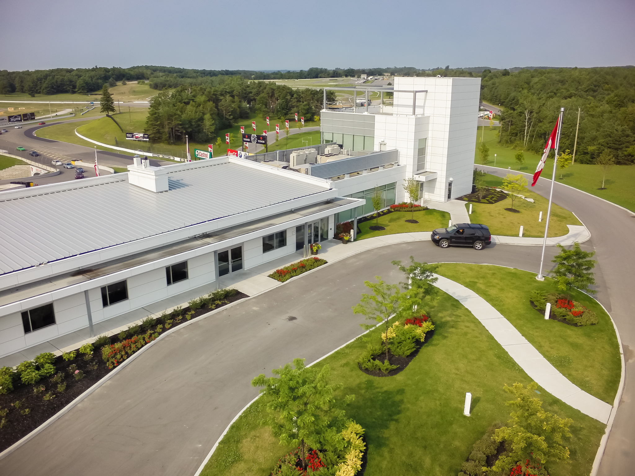 Aerial view of a white building with landscaped gardens and a driveway loop leading to the entrance, with Canadian flags along the road.