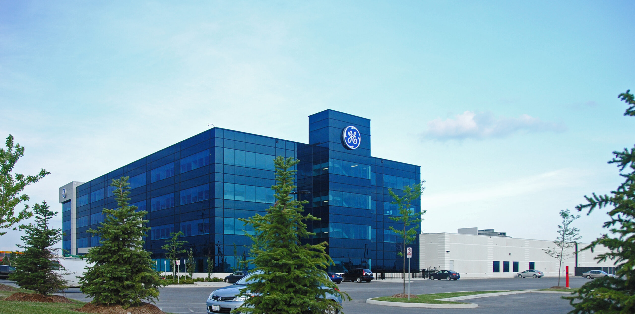 GE office complex with a deep blue glass façade, seen from a distance behind rows of young trees.