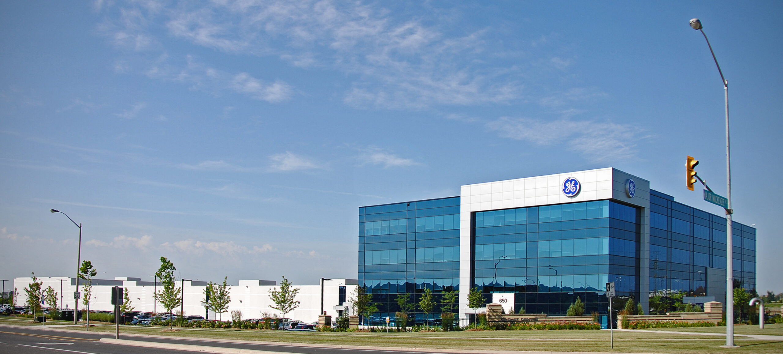 Panoramic street-side view of GE’s blue glass building with adjacent white industrial structures.