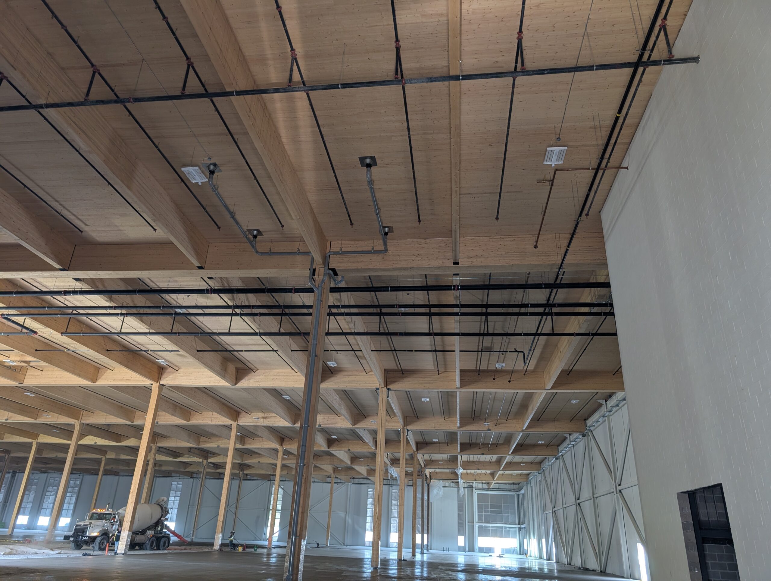 Interior of a mass timber facility with exposed wooden beams, suspended piping and utilities, and active concrete finishing work taking place on the floor.