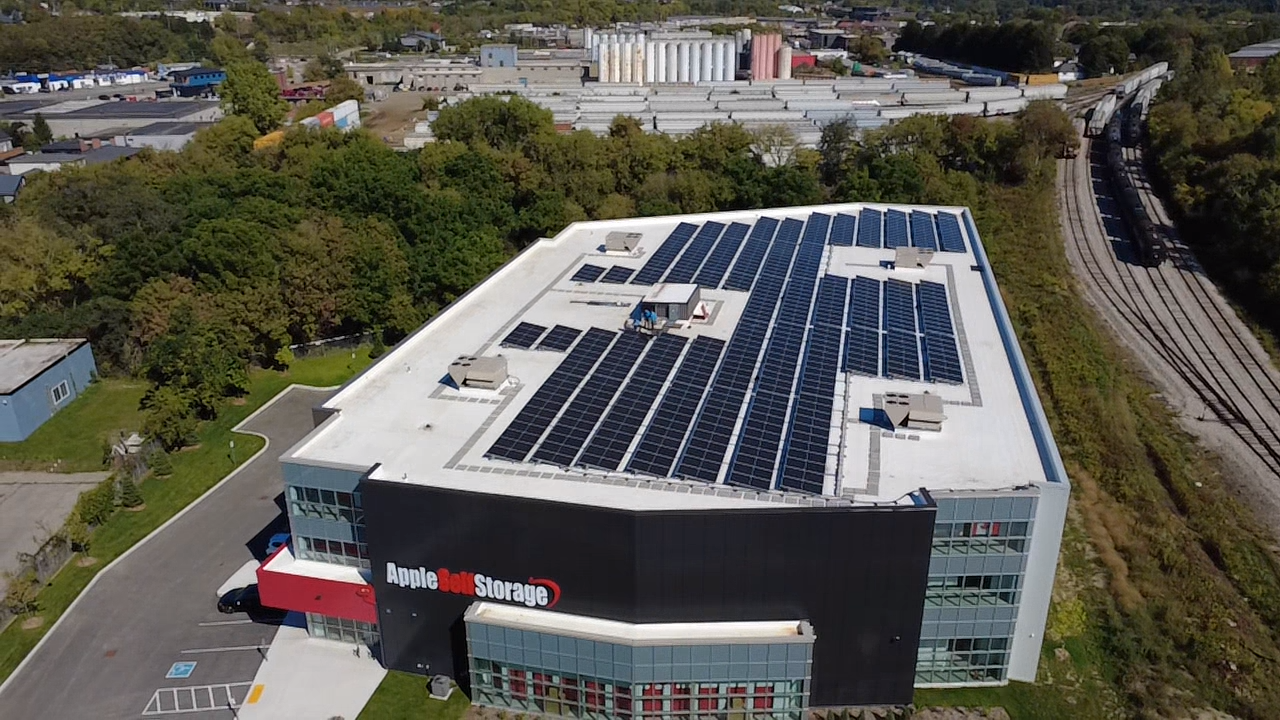 Aerial view of an Apple Self Storage facility with solar panels covering the roof, surrounded by trees and nearby roads.