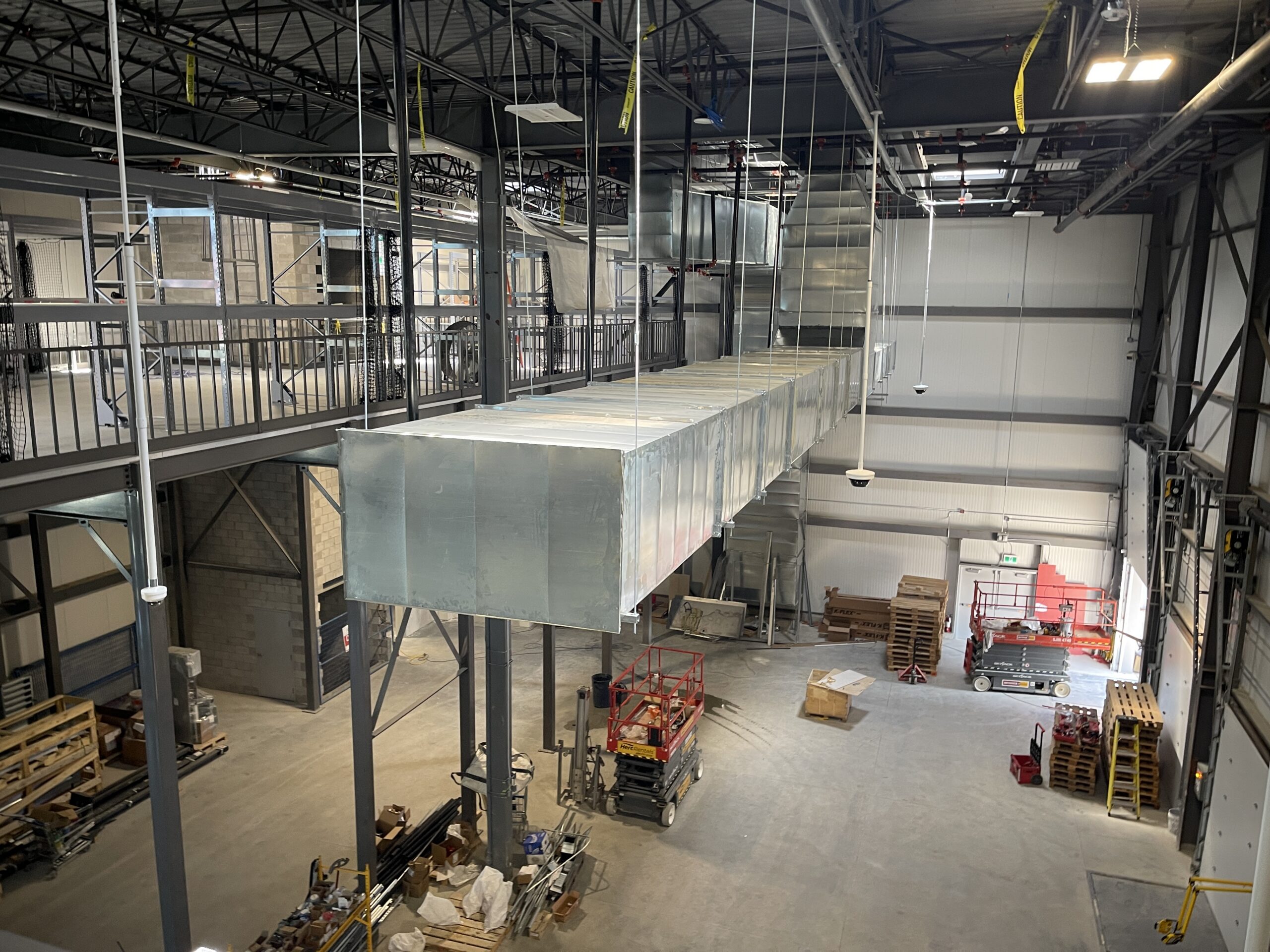 Interior of an industrial facility showing overhead HVAC ductwork, steel structure, mezzanine walkways, and construction equipment on the warehouse floor.