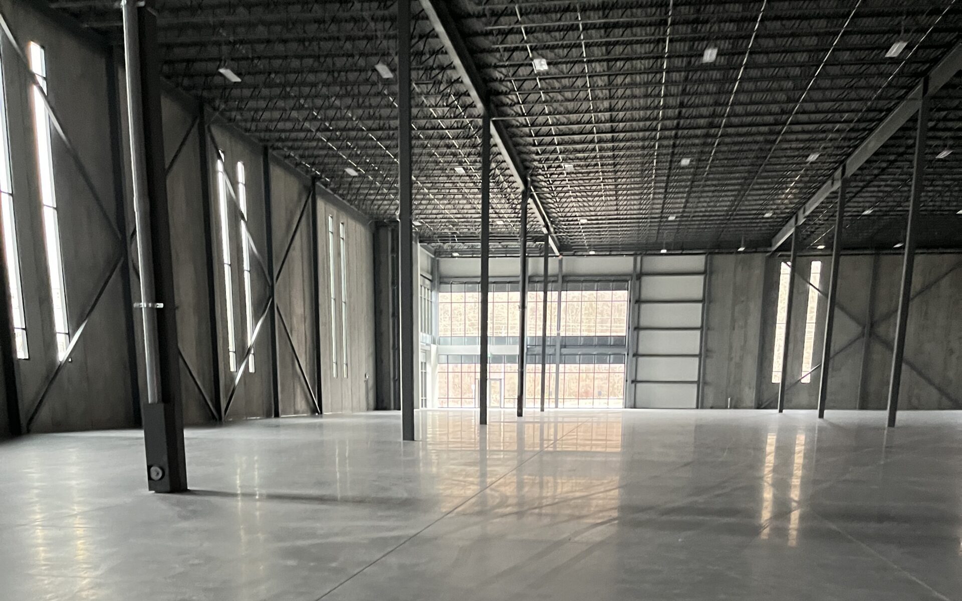 Interior view of a large industrial warehouse with polished concrete floors, structural steel columns, high ceilings, and natural light from perimeter windows.